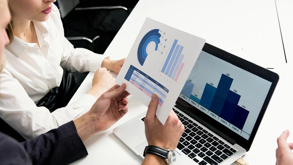 Two people discussing printed charts and laptop data in a meeting